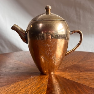 May include: A vintage brass teapot with a lid and handle. The teapot has an etched design on the side, featuring a landscape scene. The teapot is sitting on a wooden table.