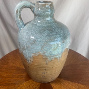 May include: A ceramic jug with a rounded body and a handle. The top half is a light blue with a crackle glaze, transitioning to a tan base. The jug sits on a wooden surface.