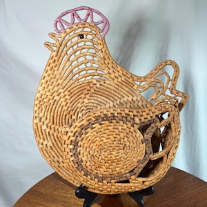 May include: A woven basket in the shape of a hen. The basket is made of natural-colored woven material with a pink comb and a dark brown eye. The hen is displayed on a black stand, showcasing its intricate design.