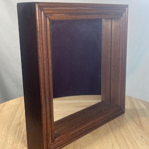 Pode incluir: Uma moldura retangular de madeira com acabamento castanho escuro. A moldura tem uma área rebaixada com fundo roxo escuro. A moldura está sobre uma superfície de madeira castanho claro.