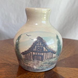 May include: A small, beige ceramic vase featuring a painted landscape. The scene shows a barn with a thatched roof, set against trees and a blue sky. The vase has a rounded body and a narrow neck.