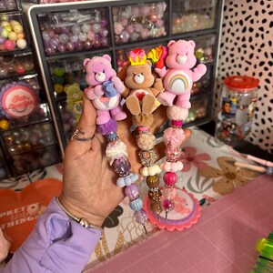 May include: Three Care Bear-themed beaded charms. Each charm features a different Care Bear character with colorful beads, ribbons, and decorative elements. The charms are held in a hand, with a craft supply storage unit in the background.