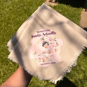 May include: A beige linen napkin with frayed edges. The napkin features a printed design with the text "MI BAUTIZO Yaretzi Antonella" and "AGOSTO 3, 2025". The design includes a watercolor illustration of a young girl surrounded by flowers.