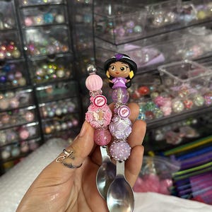 以下が含まれることがあります： プリンセスキャラクターがついた装飾的なスプーンが2本。スプーンはピンク、パープル、ホワイトのビーズと花で飾られています。プリンセスキャラクターはパープルのドレスを着て、茶色の髪をしています。