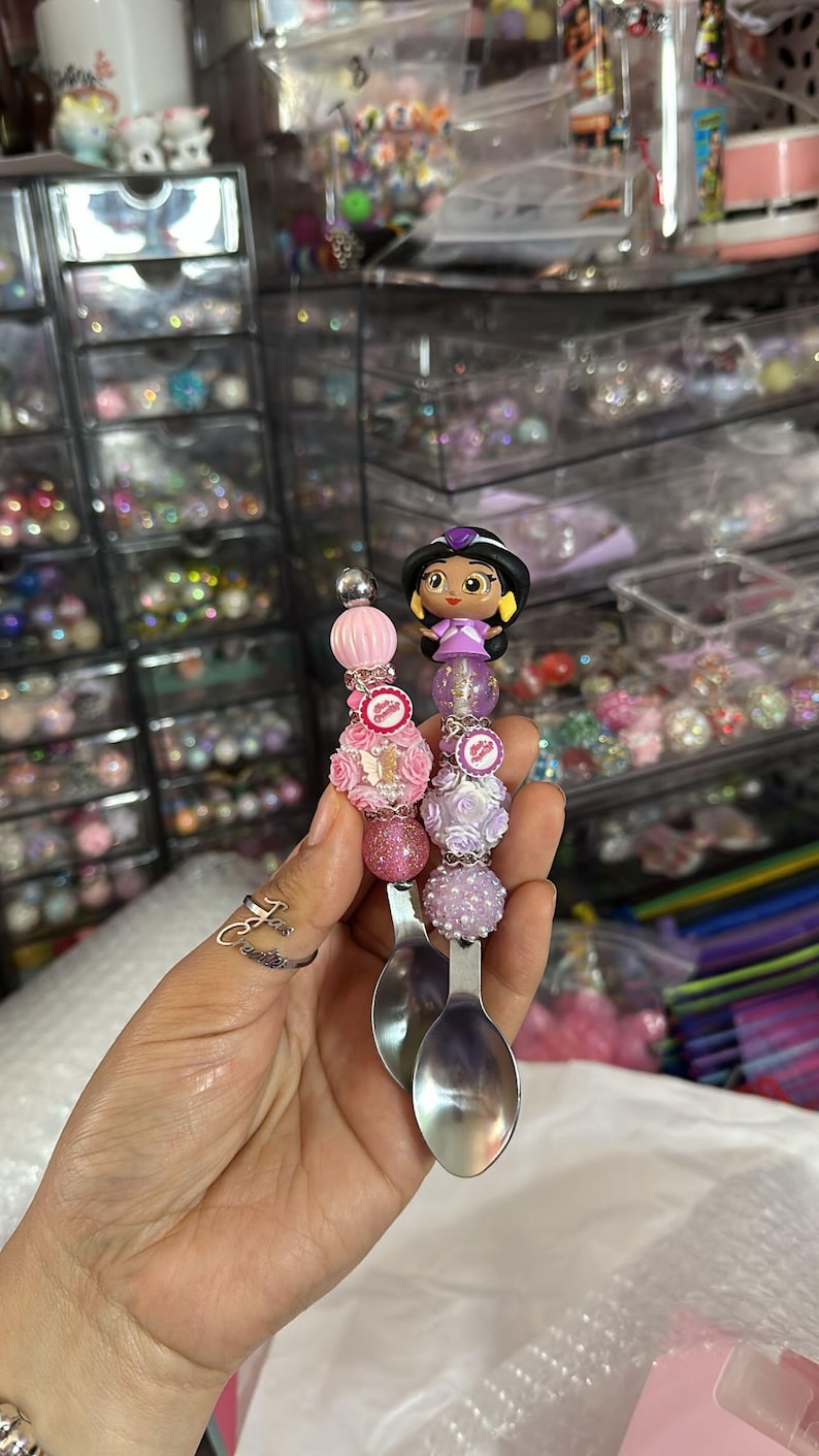 以下が含まれることがあります： プリンセスキャラクターがついた装飾的なスプーンが2本。スプーンはピンク、パープル、ホワイトのビーズと花で飾られています。プリンセスキャラクターはパープルのドレスを着て、茶色の髪をしています。
