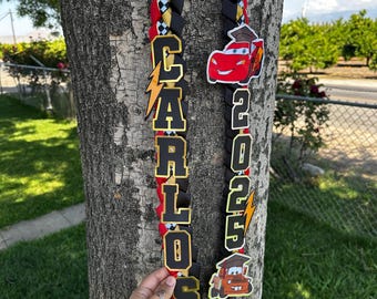 May include: A graduation lei with a red, black, and gold braided design. The lei features the name "CARLOS" in gold letters and the year "2023". Decorative Lightning McQueen and Mater car characters are also included.