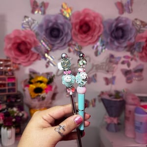 以下が含まれることがあります： 銀色のトップとカラフルなビーズのアクセントが付いたターコイズブルーのペンが2本。ペンは銀色の金属製の先端を持ち、小さなチャームで飾られています。チャームには「Topper」と「Mark」と書かれています。