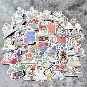 Puede incluir: Un conjunto de pegatinas que representan libros abiertos con arreglos florales coloridos. Algunas pegatinas tienen el texto "Teacher". Las pegatinas son de varios colores, incluyendo rojo, rosa, azul y amarillo, y están dispersas sobre una superficie gris.