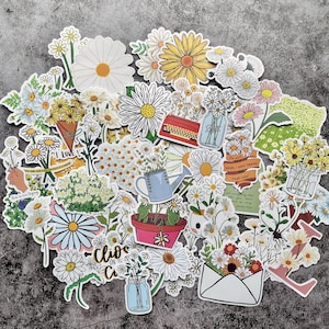 Könnte beinhalten: Eine Sammlung von Gänseblümchen-Aufklebern in verschiedenen Designs. Die Aufkleber zeigen weiße, gelbe und rosa Gänseblümchen sowie Illustrationen von Vasen, Gießkannen und Text. Die Aufkleber sind auf einer grauen Oberfläche angeordnet.