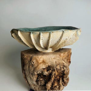 May include: A handcrafted ceramic bowl with a textured, cream-colored exterior and a teal interior. The bowl has a unique, fluted design and sits atop a rustic wooden base, showcasing its artisanal quality.