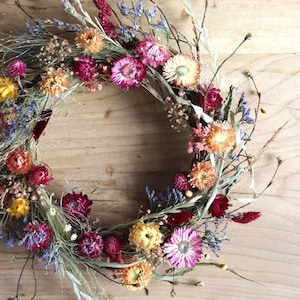 Puede incluir: Una corona de flores secas con una variedad de colores, incluyendo rosa, morado, naranja y amarillo. La corona está hecha con materiales naturales y es perfecta para agregar un toque de encanto rústico a cualquier hogar.