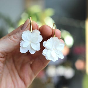 Può includere: Un paio di orecchini a fiore bianchi con ganci color oro. Gli orecchini presentano delicati petali a strati, creando un design floreale tridimensionale. Gli orecchini sono tenuti contro uno sfondo sfocato.