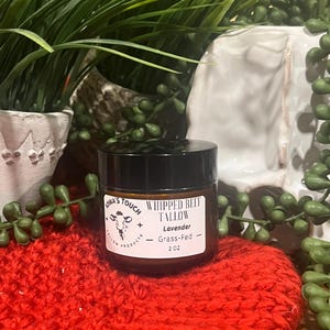 Puede incluir: Un pequeño frasco de vidrio con tapa negra que contiene sebo de res batido con aroma a lavanda. El frasco está etiquetado con "Whipped Beef Tallow - Lavender - Grass-Fed - 2 oz" y tiene un logotipo de silueta de vaca. El frasco está sobre una superficie de punto rojo.