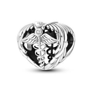 May include: A silver heart-shaped charm featuring a central caduceus symbol with wings and a small embedded clear stone. The metal surface is engraved with the text S925 at the top, indicating sterling silver composition in this jewelry piece.
