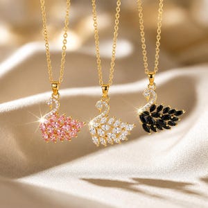 Può includere: Tre collane color oro con ciondoli a forma di cigno. Ogni ciondolo è incastonato con piccole pietre chiare lungo il collo e pietre taglio marquise che formano le ali, disponibili nelle varianti di colore rosa, trasparente e nero.