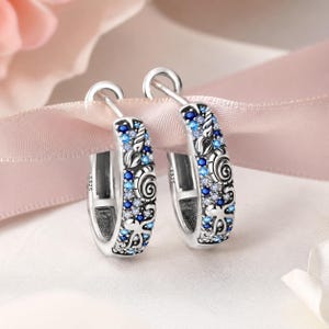 May include: A pair of sterling silver hoop earrings featuring an intricate ocean-themed design with embossed seashells and starfish. The surface is embellished with a gradient of blue and clear cubic zirconia stones and includes an S925 hallmark stamp.