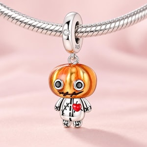Può includere: Un ciondolo in argento a forma di figura con testa di zucca, occhi neri circolari, bocca cucita e un cuore rosso sul petto. Il gancio reca il marchio S925 e include una piccola pietra trasparente. Il ciondolo è appeso a una catena d'argento.