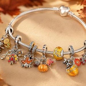 Könnte beinhalten: Ein silbernes Schlangenketten-Charm-Armband mit herzförmigem Verschluss und verschiedenen herbstlichen Anhängern, darunter eine Sonnenblume, ein Baum mit roten und orangen Blättern, ein Ahornblatt, ein Kürbis, eine Biene, ein Honigtopf mit der Aufschrift HONEY und eine Eichel.