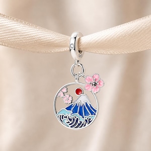 May include: A sterling silver charm featuring a circular frame with a blue and white mountain, a red sun, and stylized waves. A small pink cherry blossom flower is attached to the top right. The bail is stamped with S925 and a small clear stone.