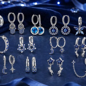 Puede incluir: Una colección de pendientes de aro plateados con varios detalles de cristales azules y transparentes. Los diseños incluyen manos de hamsa, ojos turcos, mariposas, estrellas, lunas y siluetas de gatos, con algunos pares que presentan colgantes.