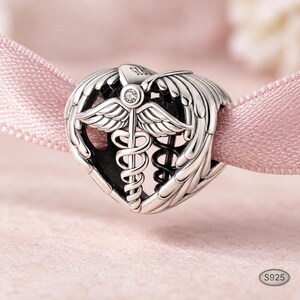 May include: A silver heart-shaped charm bead featuring a caduceus symbol with wings and a small clear stone at the top. The metal surface is engraved with S925. The charm has an openwork design with dark oxidized detailing in the recessed areas.