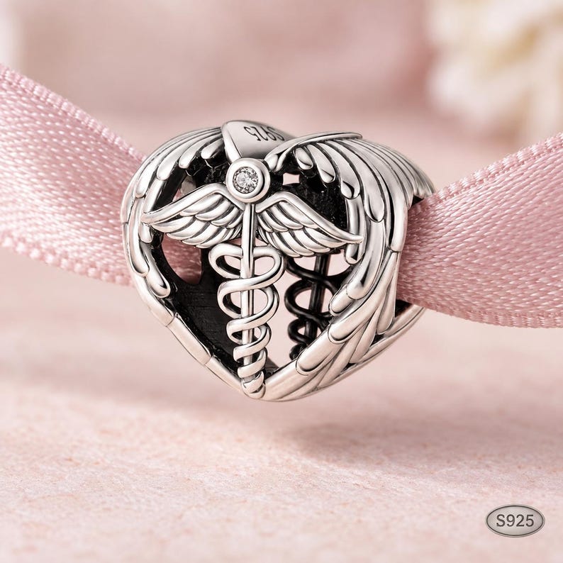 May include: A silver heart-shaped charm bead featuring a caduceus symbol with wings and a small clear stone at the top. The metal surface is engraved with S925. The charm has an openwork design with dark oxidized detailing in the recessed areas.