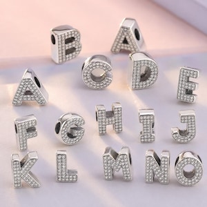 Puede incluir: Una colección de dijes con letras del alfabeto en metal plateado, cada uno incrustado con numerosas piedras pequeñas, claras y brillantes. Se muestran las letras de la A a la O, con un orificio central para enhebrar en una cadena o pulsera.
