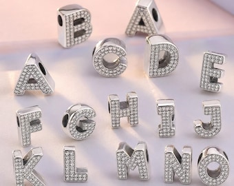 May include: A collection of silver-toned metal alphabet letter charms, each encrusted with numerous small, clear, sparkling stones. The letters A through O are displayed, featuring a hollow center hole for threading onto a bracelet or necklace chain.
