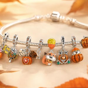 Può includere: Un bracciale a catena snake in argento con molteplici ciondoli a tema autunnale, tra cui uno scoiattolo, zucche, un corno dell'abbondanza e una carrozza. I ciondoli sono decorati con smalto arancione, giallo e verde, cristalli e la scritta HAPPY.