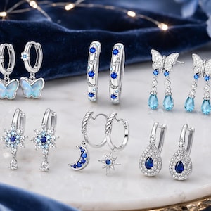 May include: A collection of six pairs of silver-toned earrings featuring various designs including iridescent butterfly charms, blue and white celestial motifs, starburst shapes, crescent moons, and teardrop-shaped blue stones accented with clear crystals.