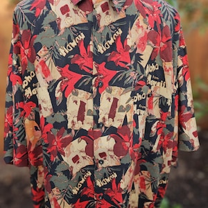 May include: A short-sleeved button-down shirt with a vibrant Hawaiian floral print. The shirt features a black background with red hibiscus flowers, green leaves, and the word "Hawaii" in black. The shirt has a collared neckline and buttoned front.