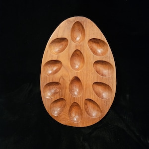 May include: A wooden egg-shaped serving tray with 12 egg-shaped indentations. The tray is made of light brown wood with visible grain patterns. The egg holder is designed to hold a dozen eggs.