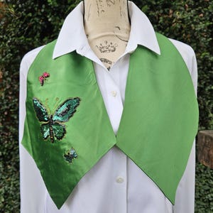 May include: A green satin vest with a butterfly design. The butterfly is embellished with sequins in shades of green and black. The vest is worn over a white collared shirt.