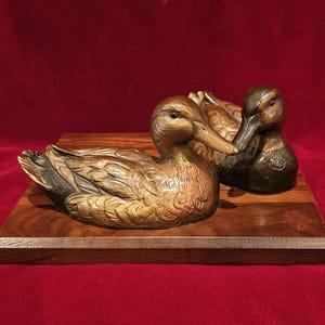 May include: Two detailed duck sculptures, one bronze and one brown, rest on a wooden base. The ducks are positioned side-by-side, showcasing intricate feather details. The base is a rectangular shape, and the background is a deep red.