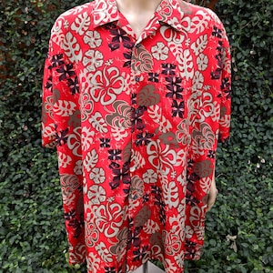 May include: A short-sleeved button-down shirt in a vibrant red color featuring a repeating tropical pattern of beige hibiscus flowers and dark brown monstera leaves. The shirt has a classic collar, a front button placket, and a chest pocket.
