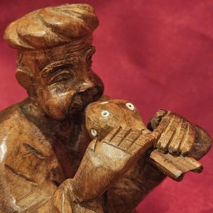 May include: A detailed wooden carving of a musician playing a wind instrument. The figure is dressed in a hat and appears to be blowing into the instrument. The carving is a warm brown color against a red background.