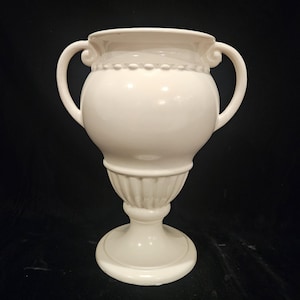 Może przedstawiać: Biały ceramiczny wazon z dwoma zakrzywionymi uchwytami. Wazon ma pękaty korpus, żłobioną podstawę i ozdobny, koralikowy brzeg. Wazon ma kremowo-biały kolor i stoi na okrągłej podstawie. Tło jest czarne.