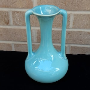 May include: A light blue ceramic vase with two curved handles. The vase has a rounded base and a flared opening. The smooth, glossy surface reflects light, highlighting its elegant shape. The vase is a decorative item.