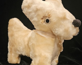 Può includere: Un giocattolo vintage a forma di cane in peluche in posizione eretta. Il cane è color crema con un naso a pompon nero e un occhio decorativo. Ha un nastro color crema intorno al collo e un abbellimento dorato vicino all'occhio.