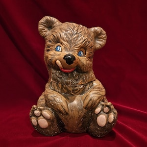May include: A brown ceramic bear cookie jar featuring a textured fur finish, blue eyes, a black nose, and a red tongue sticking out. The bear is in a seated position with visible paw pads on its feet, and the lid is integrated into the midsection.