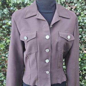 May include: A brown jacket with a collared neckline, two chest pockets, and silver buttons. The jacket has a fitted silhouette and long sleeves. The garment is displayed against a backdrop of green foliage.