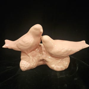 May include: A light pink ceramic bird figurine featuring two birds perched together. The birds are detailed with textured feathers and sit on a textured base. The figurine is a decorative item.