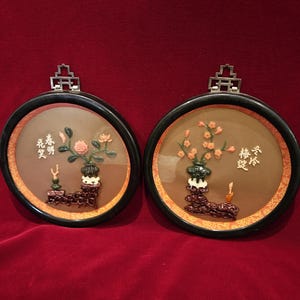 May include: Two round, framed wall art pieces with black frames and silver metal hangers. Each features a central floral arrangement in a vase, with Chinese characters. The background is a warm brown color.