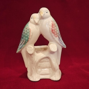 May include: A white ceramic figurine of two birds perched together. The birds have accents of green, pink, and purple on their wings. The birds are on a white base with a small opening, possibly for flowers.