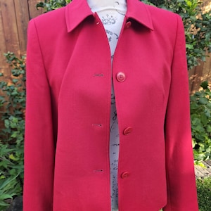 May include: A bright red blazer with a classic collar and three matching buttons. The jacket is displayed on a mannequin against a backdrop of green foliage. The blazer has long sleeves and a tailored fit.