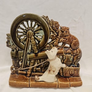 May include: A ceramic figurine depicting a dog pulling a cart with a spinning wheel. The figurine is primarily brown and cream-colored, with a detailed design of the wheel and a dog on a leash. The base is a brick-like pattern.