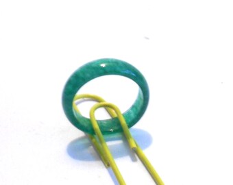 May include: A vibrant green jade ring with a smooth, polished surface. The ring is a solid band of color, showcasing the natural variations in the stone. A yellow paperclip is partially visible, adding a contrasting element.