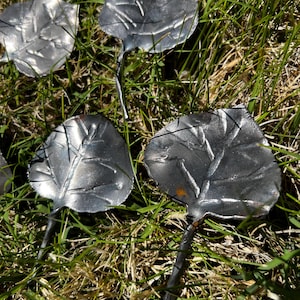 Puede incluir: Primer plano de varias estacas de jardín de hojas de metal plateado con vetas detalladas, descansando en hierba verde. Las hojas tienen un aspecto texturizado y están unidas a finos tallos, creando un elemento decorativo natural para espacios exteriores.