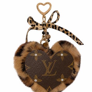 May include: A heart-shaped keychain featuring brown faux fur with a leopard print pattern. A smaller brown leather heart with the LV monogram and four small crystals is centered on the fur. It includes a gold-tone clasp and a leopard print ribbon.