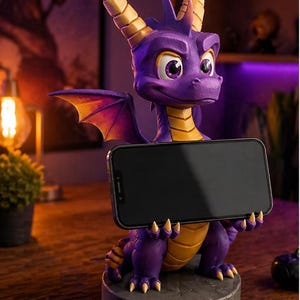Puede incluir: Una figura de dragón morado sosteniendo un teléfono inteligente negro. El dragón tiene cuernos, garras y escamas ventrales amarillas, con alas naranjas y moradas. La figura se asienta sobre una base de piedra gris, ideal para un escritorio o estante.